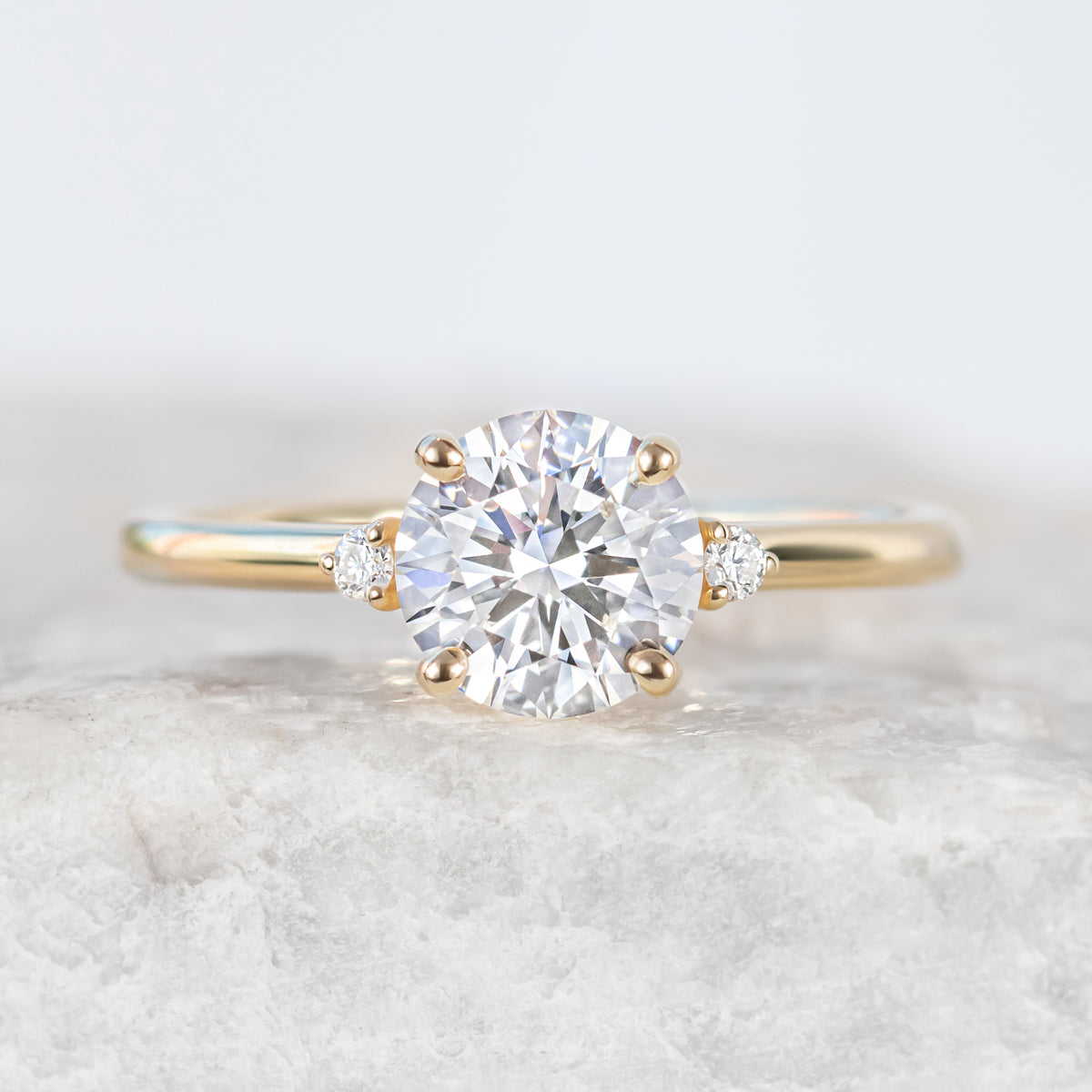 A vintage-inspired engagement ring with a brilliant round cut center diamond and two ethically sourced side stones, set in a yellow gold band.