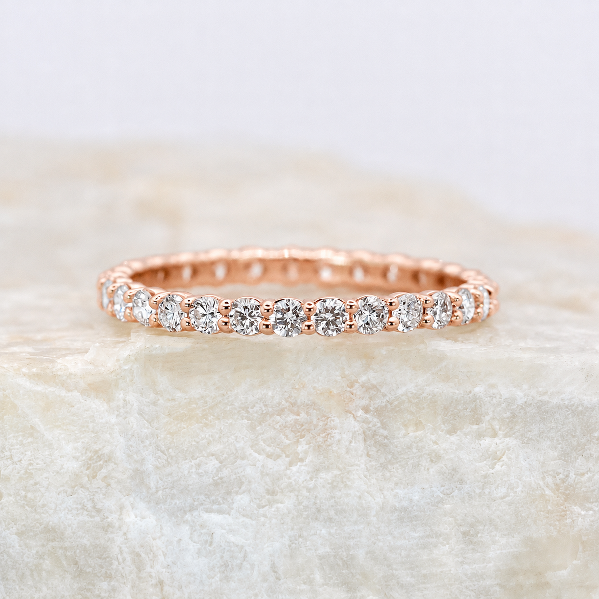 A rose gold eternity ring with round-cut clear gemstones, resting on a textured beige stone surface against a soft white background.