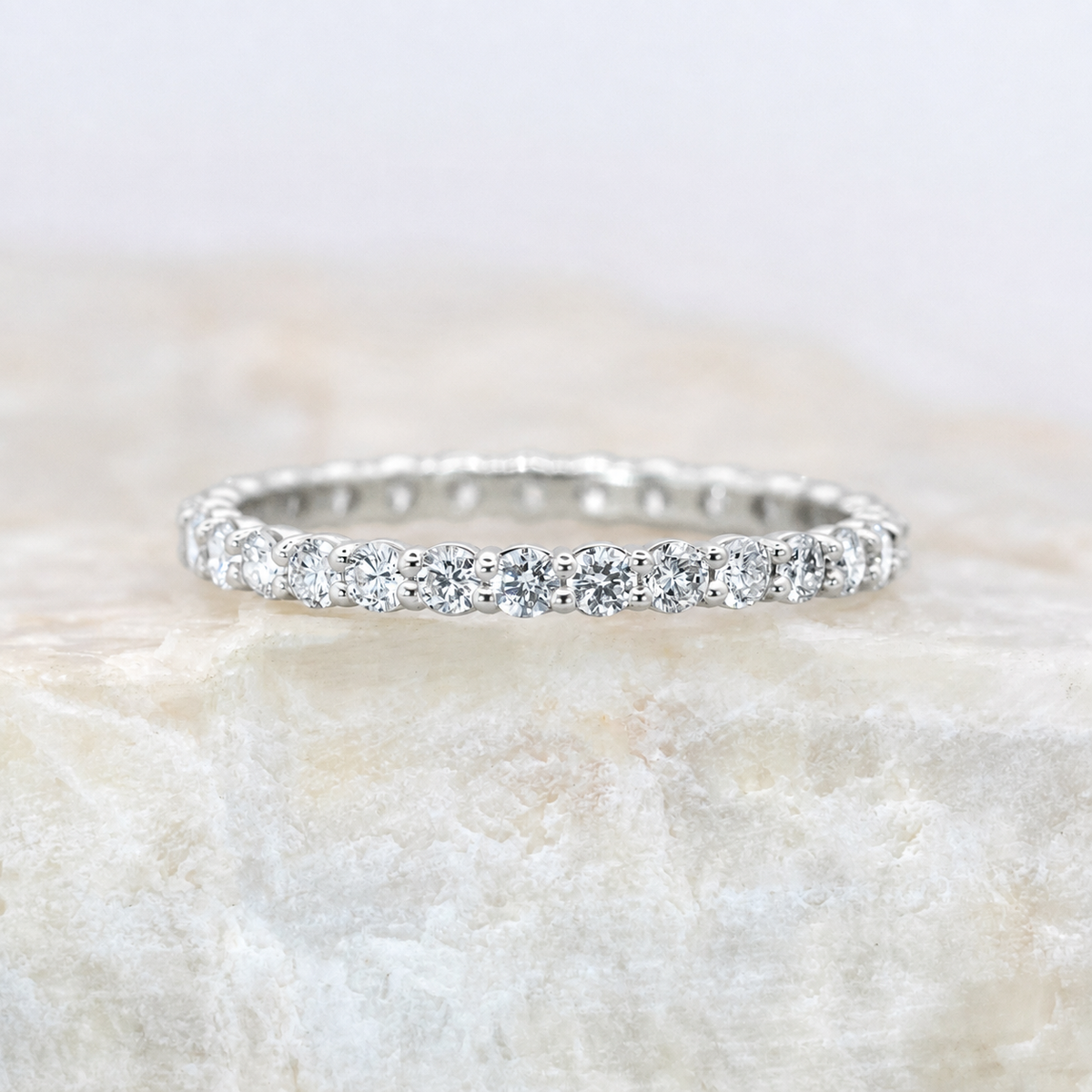 A silver diamond eternity ring set with round brilliant-cut diamonds, resting on a textured light beige stone surface.