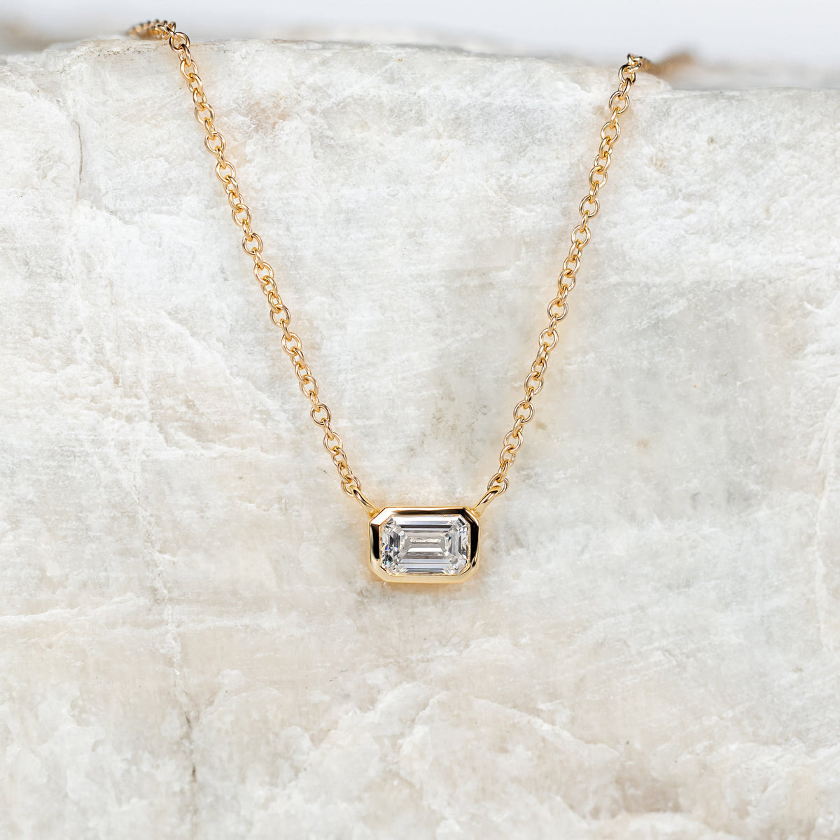 Gold necklace with a bezeled emerald cut diamond on a textured white surface