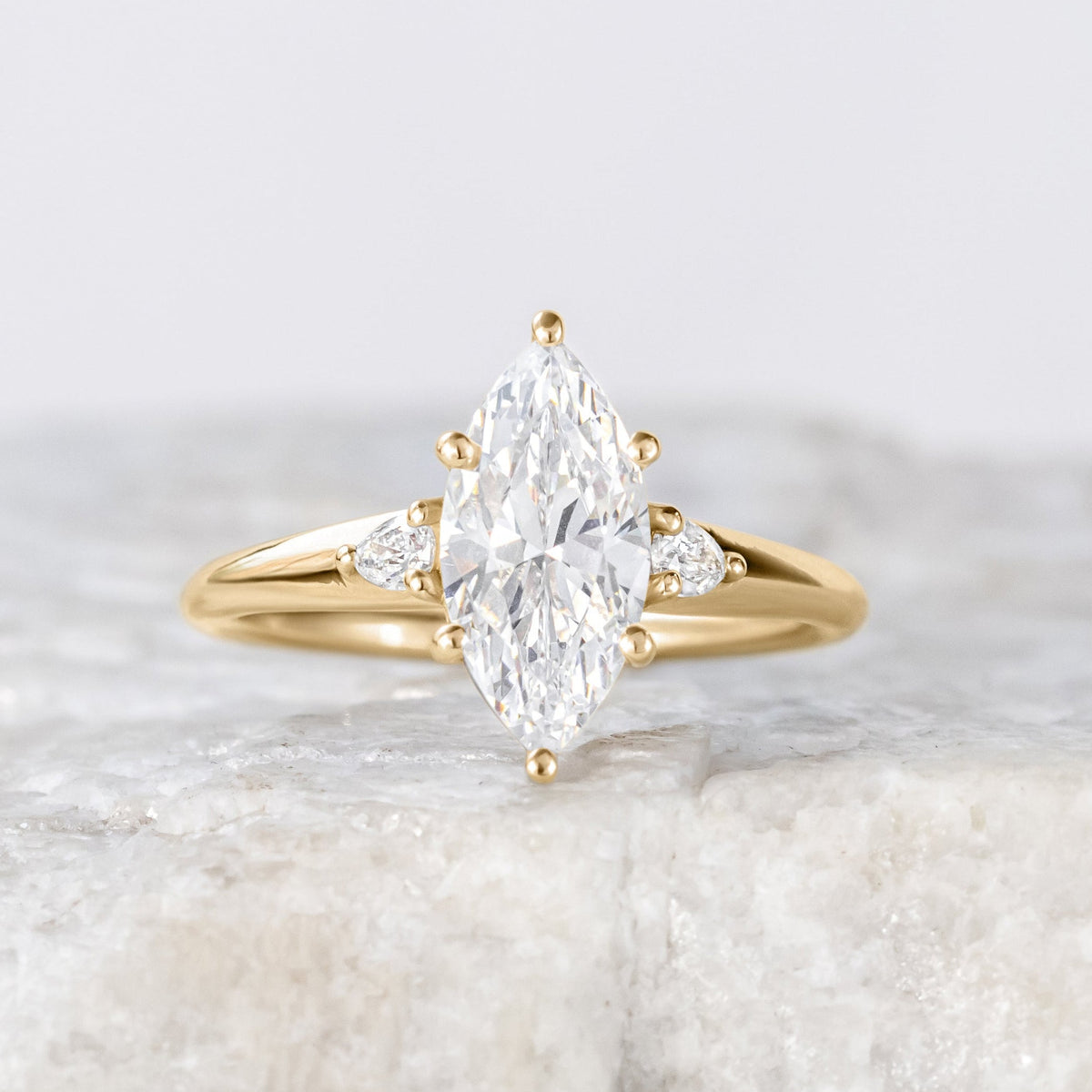 A marquise cut diamond engagement ring with pear-cut side stones set in a gold band, displayed on a marble surface.