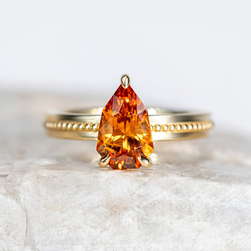 A yellow gold ring featuring a pear-cut orange gemstone, displayed on a textured white stone surface against a soft white background.