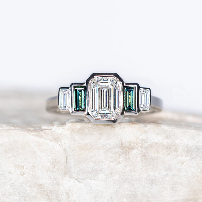 An Art Deco style silver ring featuring a central baguette-cut clear gemstone flanked by green emerald-cut side stones, displayed on a textured light beige stone block against a white background.