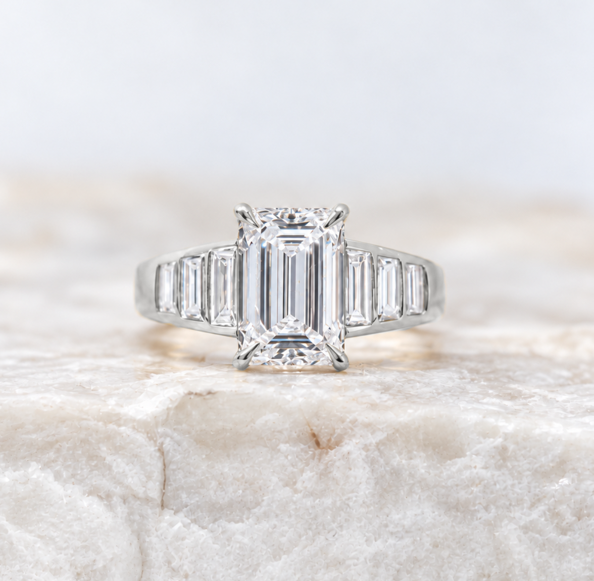 An emerald-cut diamond engagement ring with baguette side stones in a silver-colored setting, resting on a textured white stone surface.