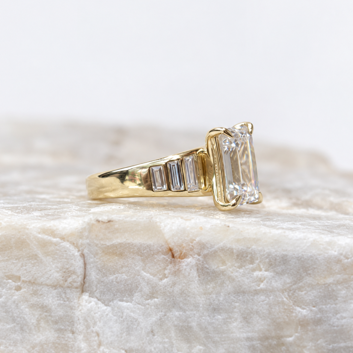 A yellow gold ring featuring a large rectangular emerald-cut clear center stone and smaller baguette-cut side stones, displayed on a textured light beige stone surface against a soft white background.