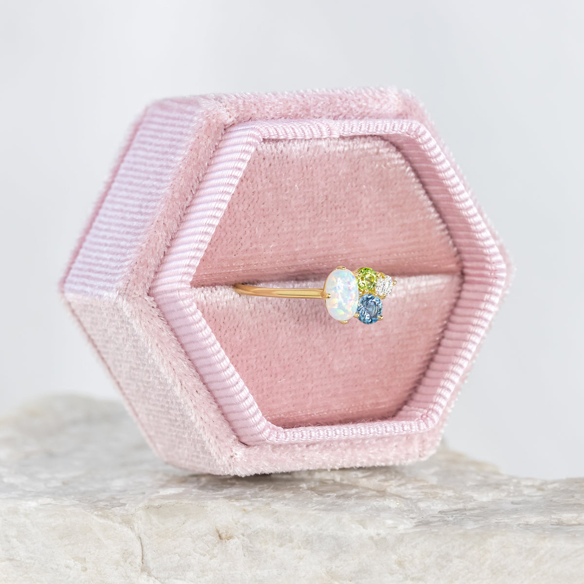 Hexagonal pink velvet ring box with a ring featuring blue and green gemstones on a light gray background.