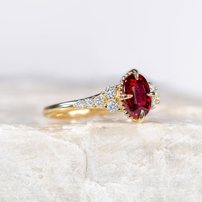 A yellow gold ring featuring an oval-cut red gemstone and a pavé setting of small clear stones, displayed on a textured white stone slab against a white background.